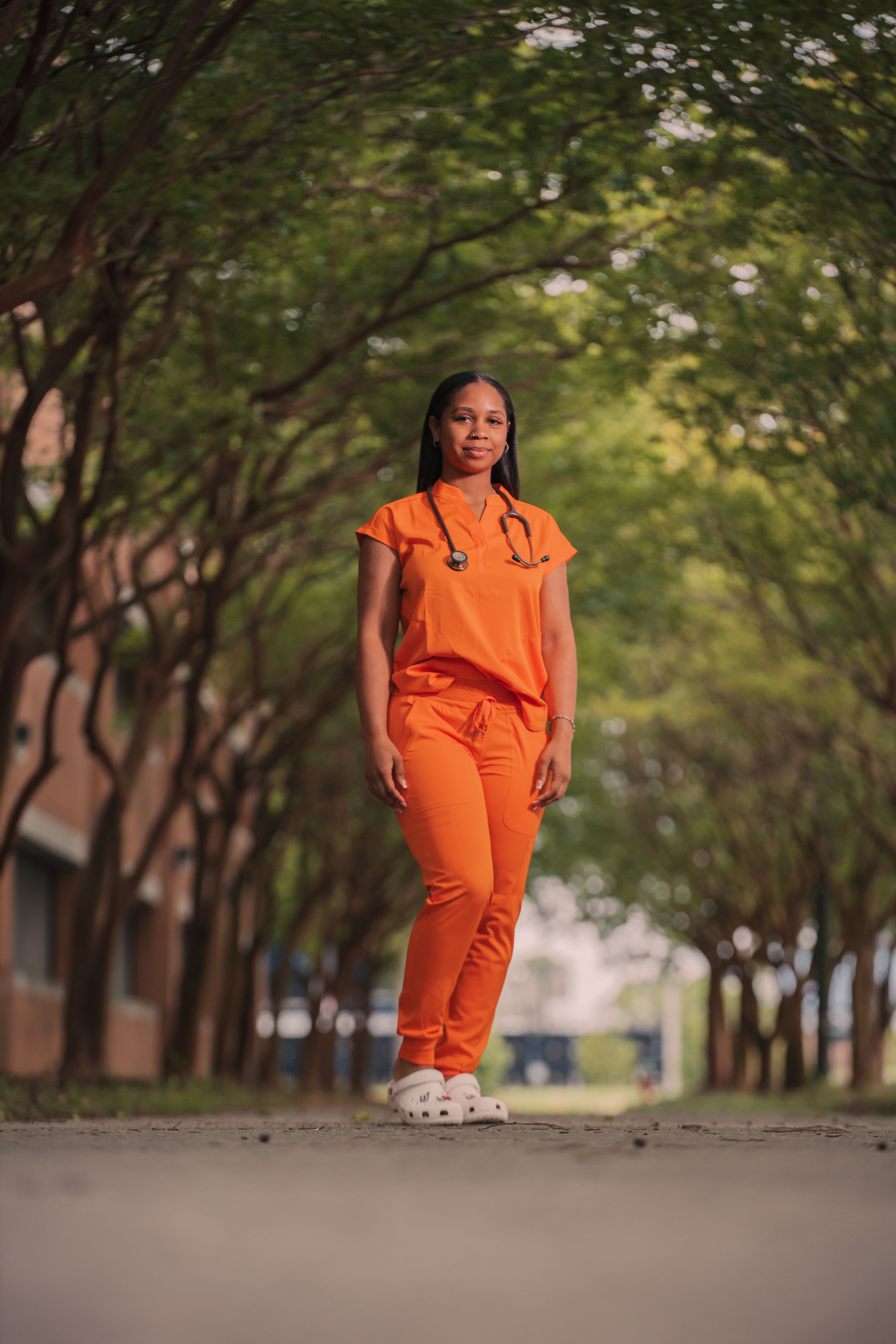 Photo of a young woman in pumpkin-colored scrubs standing under an arch of trees